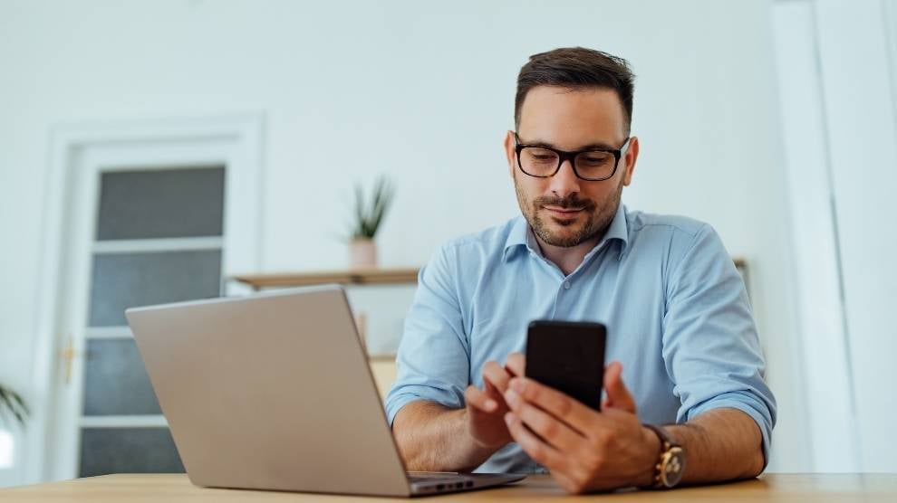 a construction marketing professional sat behind a laptop and using his phone to post on social media